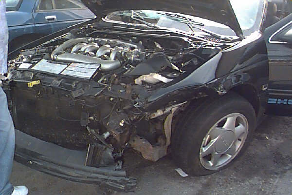 1993 Ford Taurus engine bay with front-end damage, hood open. Another car is parked to the left