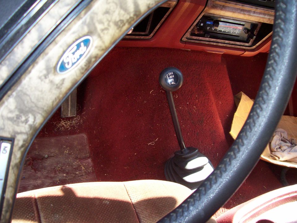 Interior shot of a 1985 Ford Bronco, showing the steering wheel with a Ford logo, the gear shifter, and the dashboard with a radio