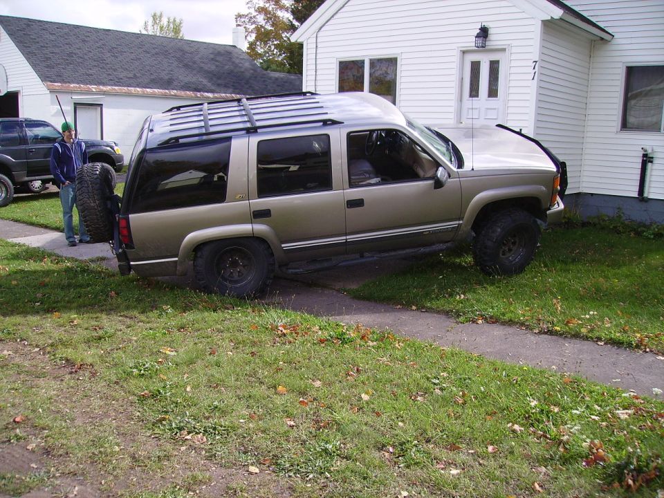 Gold Chevrolet Tahoe with roof rack and spare tire mounted on rear door, parked on grass next to a house