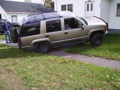 2000 Chevrolet Tahoe - A gold SUV is parked on the lawn next to a sidewalk, with a spare tire mounted on the rear.