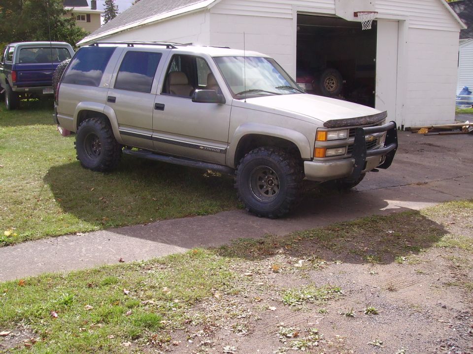 Lifted tan Chevy Tahoe with brush guard and mud tires parked beside a white block garage