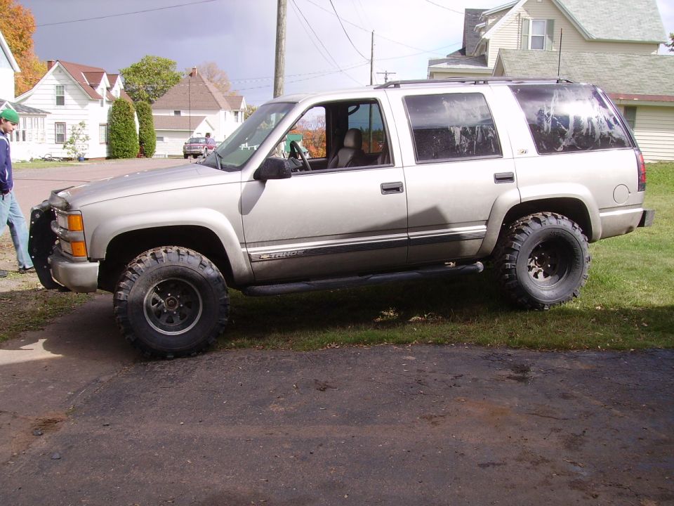 Silver lifted Chevrolet Tahoe parked on grass, side view. Has large off-road tires and a brush guard