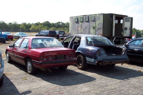 1991 Ford LTD Crown Victoria - Two sedans are parked in a lot, with a military-style trailer in the background.