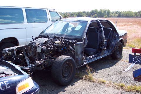 1991 Ford LTD Crown Victoria - A heavily damaged sedan sits in a lot, its engine compartment exposed and doors removed.