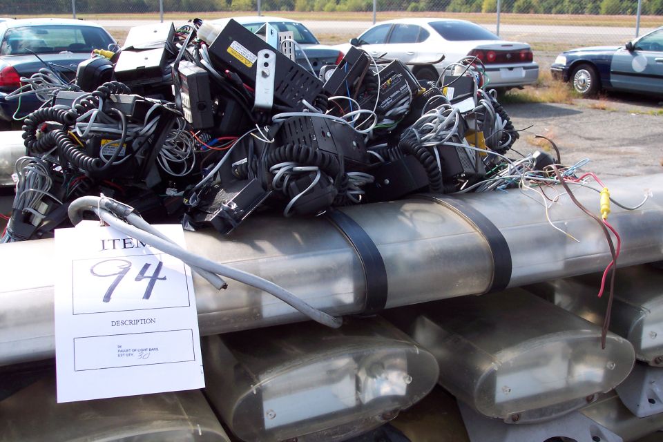 Pile of police light bars and electronic equipment, with a tag labeled 'ITEM 94' and 'PALLET OF LIGHT BARS'