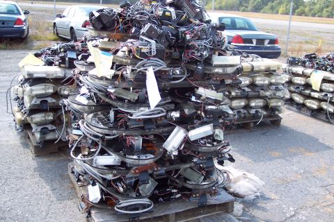 1991 Ford LTD Crown Victoria - Discarded light bars and electrical components are piled high on pallets, with two vehicles visible in the distance.