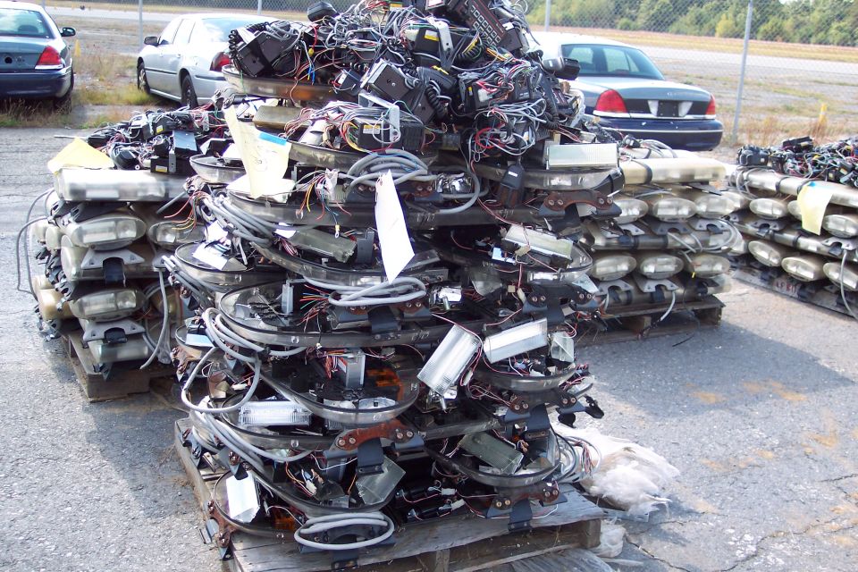 Pile of stacked light bars and wiring on wooden pallets, with two cars in the background