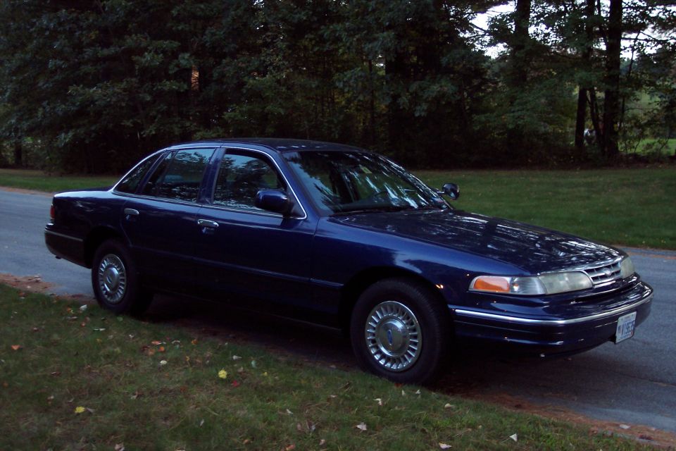 Dark blue sedan parked on grassy shoulder next to a road. Trees in background