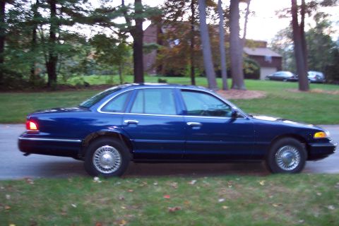 1991 Ford LTD Crown Victoria - A blue 1991 Ford Crown Victoria LTD sedan is parked on the side of a road with trees and a house in the background.