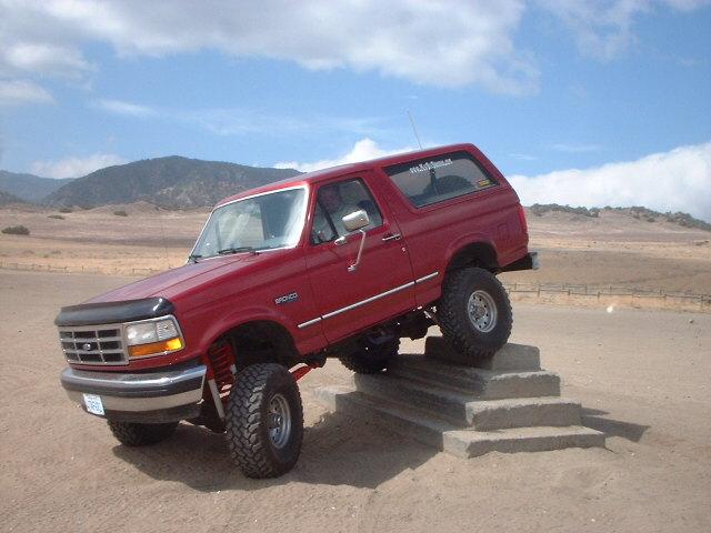 Red lifted Ford Bronco SUV climbing concrete steps in a dusty lot with mountains behind.