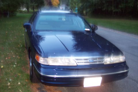 1991 Ford LTD Crown Victoria - A dark blue sedan is parked on the shoulder of a road with trees in the background.