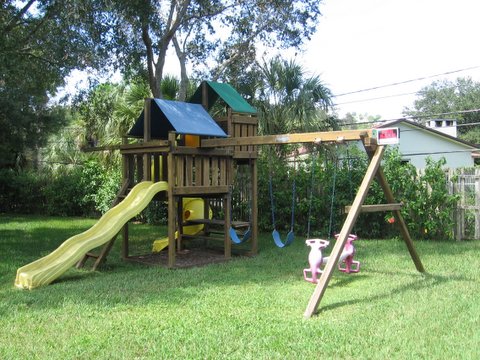 1993 Ford C-350 (Centurion Conversions) - A wooden playground set with a slide and swings is situated in a backyard with green grass and trees.