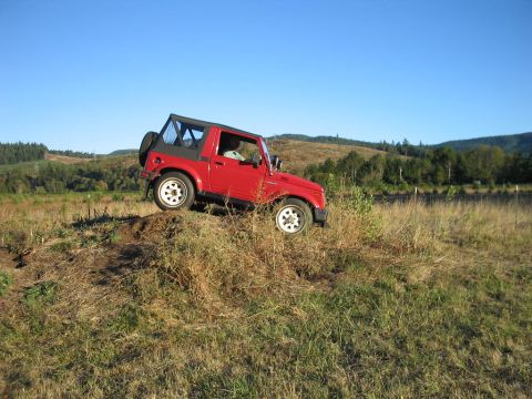 1987 Suzuki Samurai - A red Suzuki Samurai navigates a dirt path on a grassy incline with trees in the background.