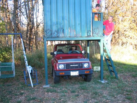 1987 Suzuki Samurai - A red 1987 Suzuki Samurai is parked under a playhouse, with two people visible in the structure above.