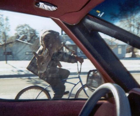 1989 Ford Bronco - A young person is seen riding a bicycle with a deer carcass draped over their shoulders, viewed from inside another vehicle.