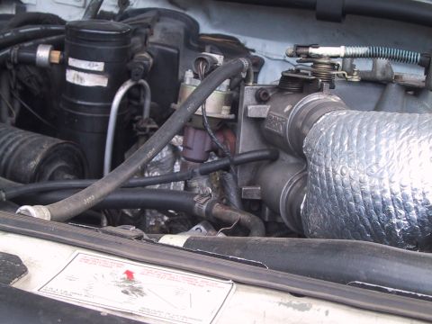 1994 Ford Bronco - Engine bay close-up of a fuel-injected V8 showing the cast throttle body with linkage and return spring, an intake tube wrapped in reflecti…