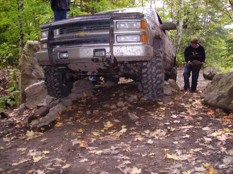 Muddy truck with a brush guard is crawling over rocks on a wooded trail. A person stands nearby