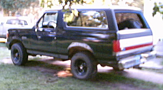 Dark lifted two-door Ford Bronco SUV viewed from rear three-quarter, parked on grass.