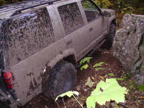 2000 Chevrolet Tahoe - Mud-covered SUV with off-road tires is stuck between rocks and mud on a trail.