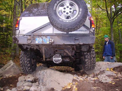 2000 Chevrolet Tahoe - This modified Chevrolet SUV is tackling a challenging off-road course with large rocks and trees surrounding it.