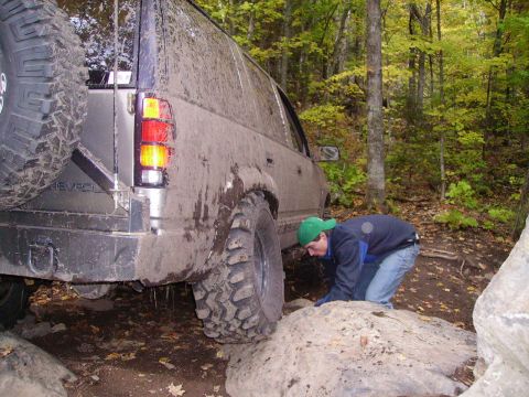 2000 Chevrolet Tahoe - A person inspects the tire of a muddy SUV navigating a rocky trail in a forest.