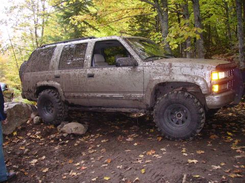 2000 Chevrolet Tahoe - This heavily muddied SUV navigates a rocky, wooded trail during autumn. Another vehicle is partially visible in the background.
