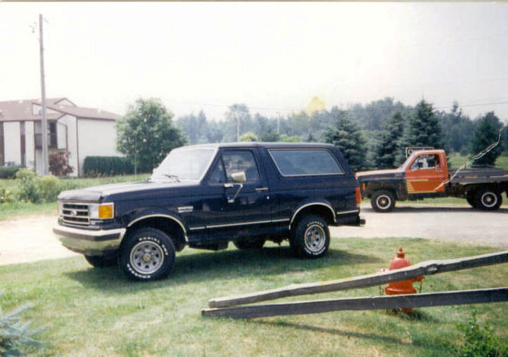 Dark blue Ford Bronco SUV side view on grass with orange flatbed truck and fire hydrant behind
