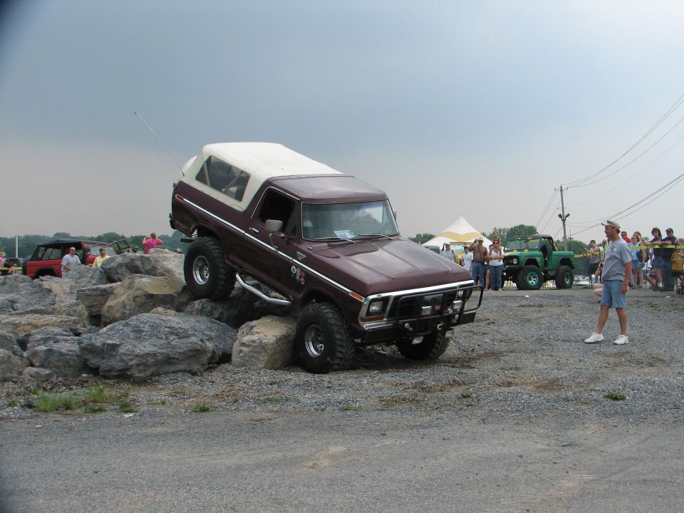 Maroon Ford Bronco with white top climbing over large rocks, green Jeep in background
