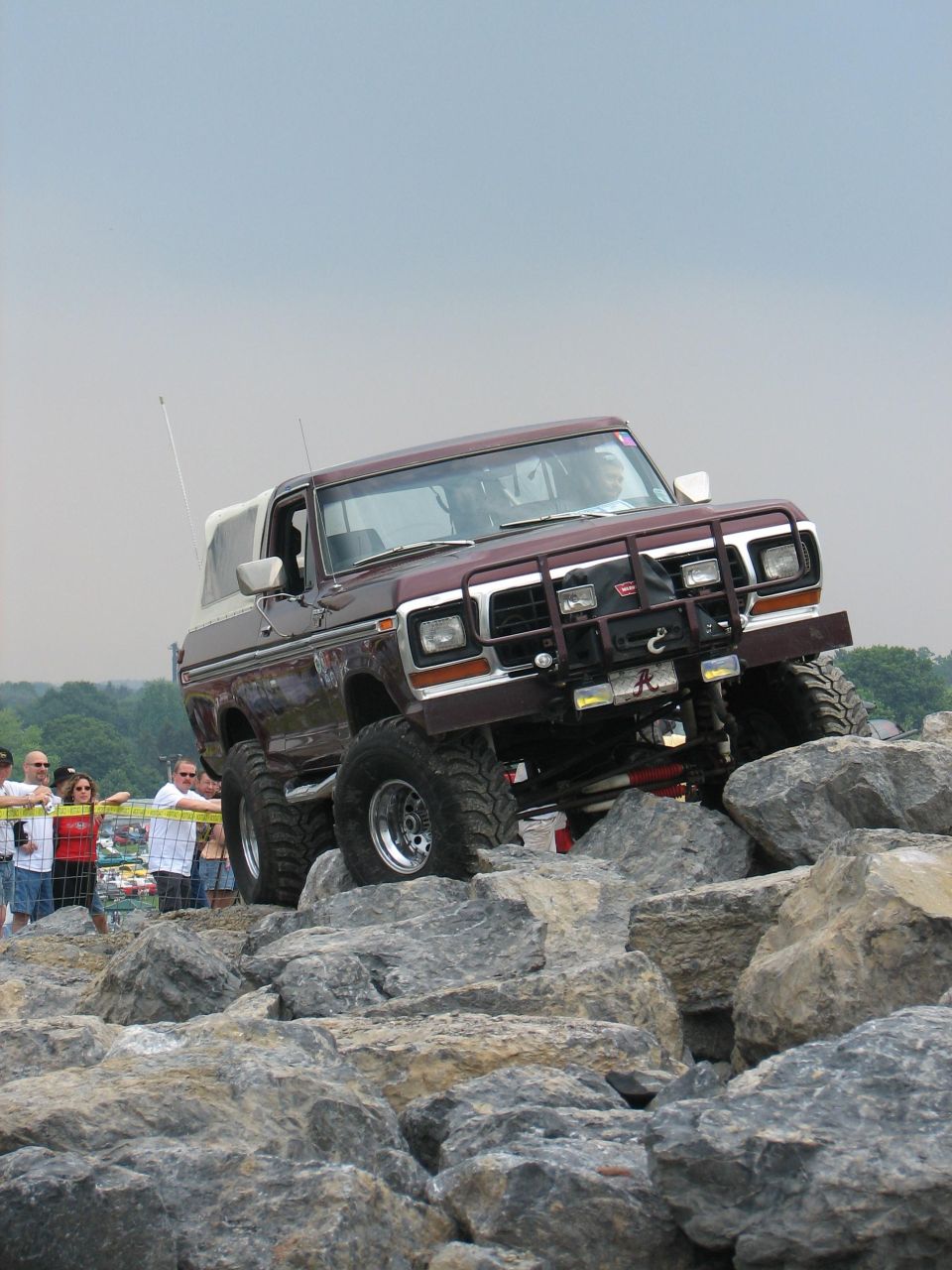 Brown 1978 Ford Bronco with a white top, lifted suspension, and bull bar, navigating a rocky terrain