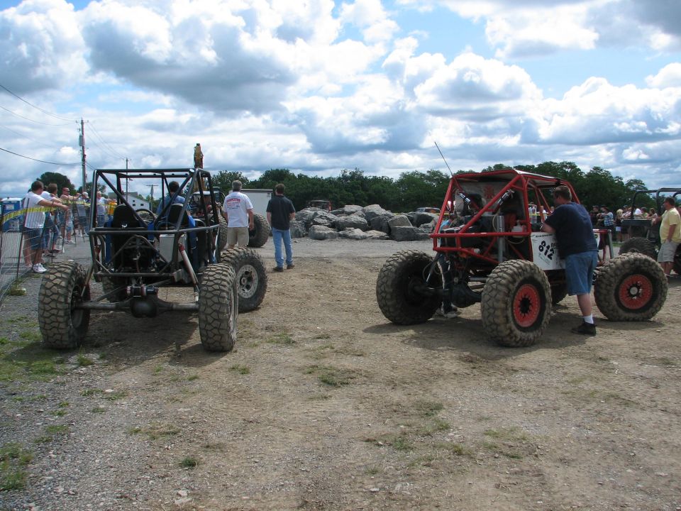 Two custom off-road buggies parked on a dirt lot, one red and one black. People are standing around them