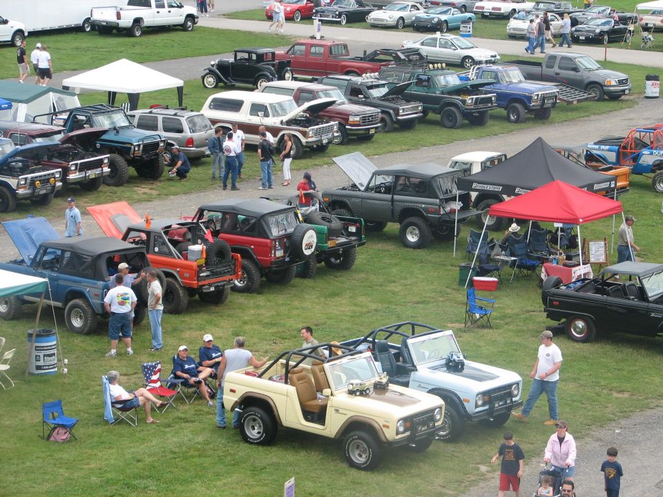 Several Ford Broncos parked on grass, some with hoods open. People milling around
