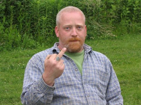 1979 Ford F-150 - Portrait of a bearded person in a blue plaid shirt and green tee standing on a grassy lawn with tall green brush behind them, making a hand…