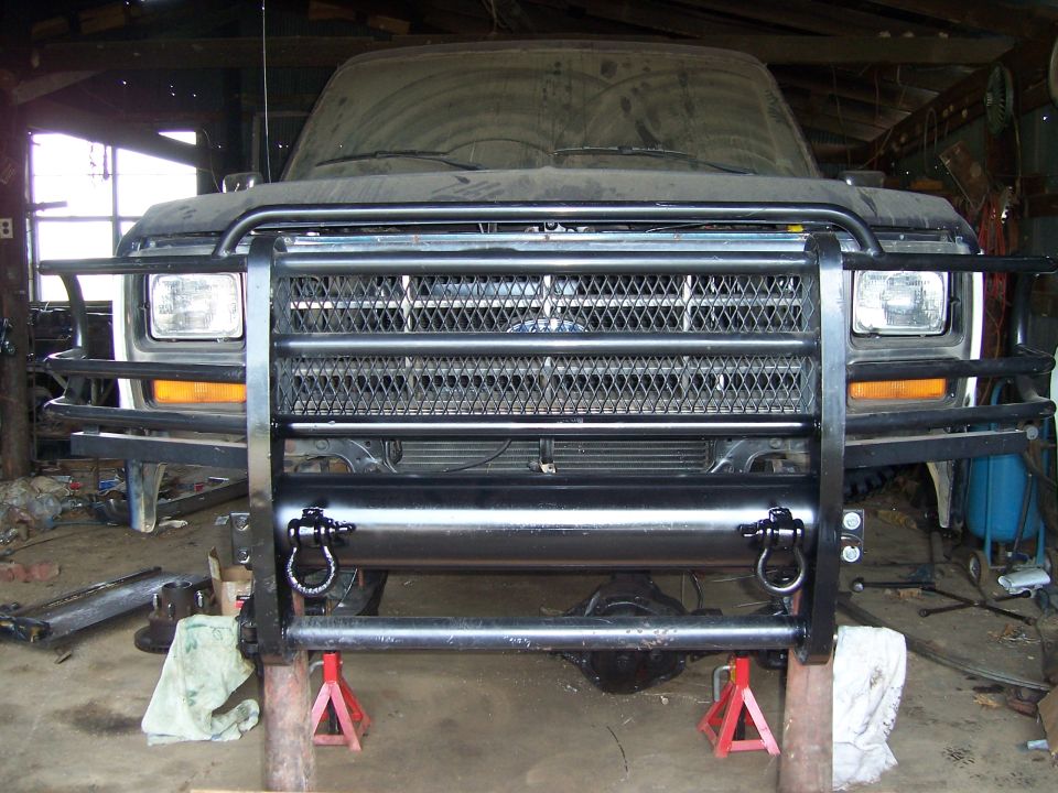 Front view of a dusty, dark-colored Ford Bronco with a large black brush guard and tow rings, supported by red jack stands