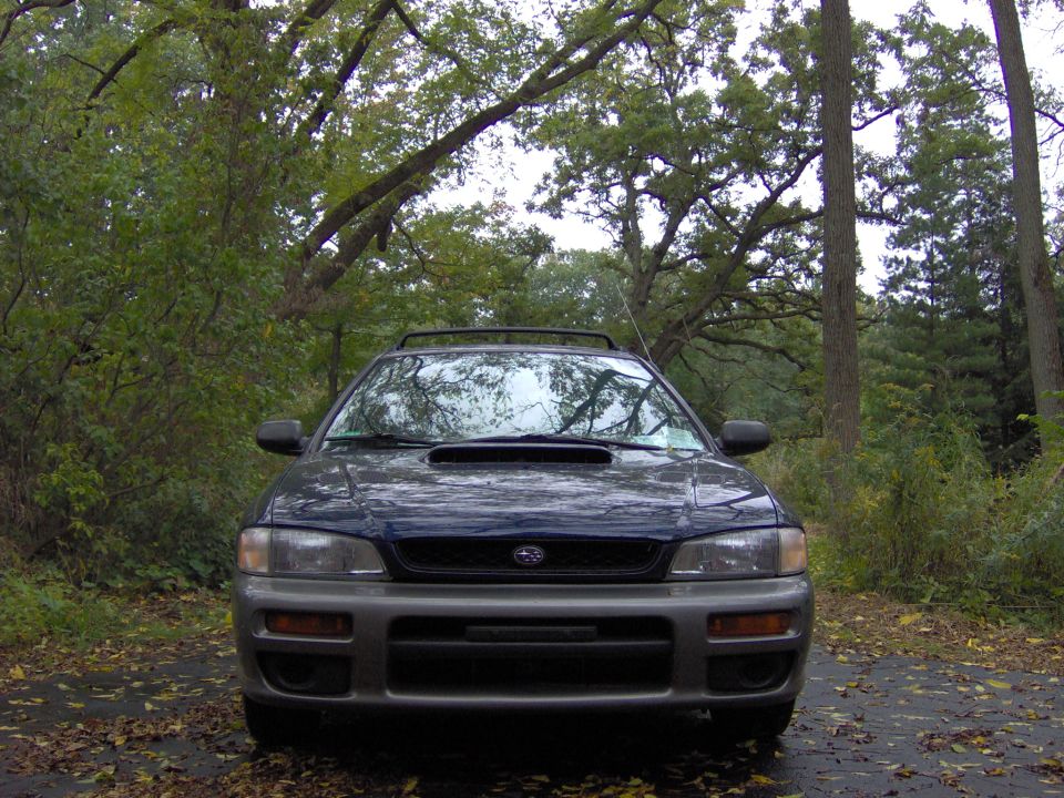 Front view of a dark blue Subaru Impreza wagon with hood scoop on a wet leafy road