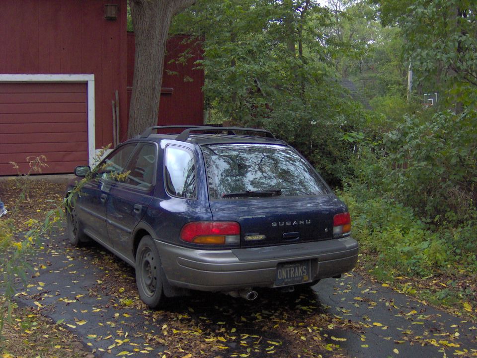 Dark blue Subaru Impreza Outback wagon parked rear-left view by red barn on leafy driveway