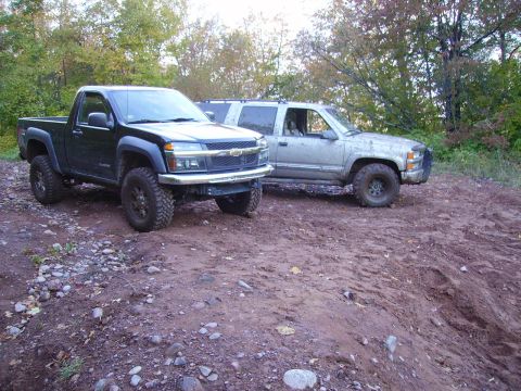 2000 Chevrolet Tahoe - Two modified trucks, a black Chevrolet Colorado and a silver Chevrolet Suburban, are parked on a dirt road with trees in the background.