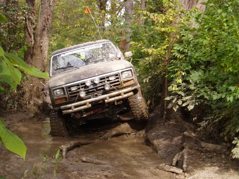 1979 Ford Bronco - A muddy 1980s Ford Bronco drives through a challenging off-road course with dense foliage.
