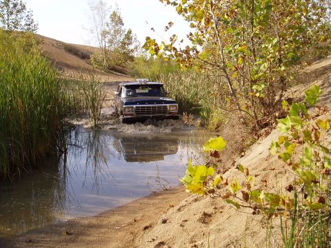 1979 Ford Bronco - Ford Bronco navigates a water crossing on a dirt trail, kicking up spray.