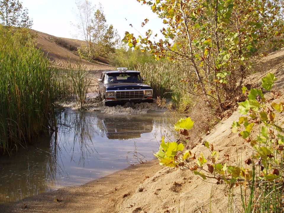 Dark blue Ford Bronco driving through a muddy water obstacle. Grille indicates 1978 model year