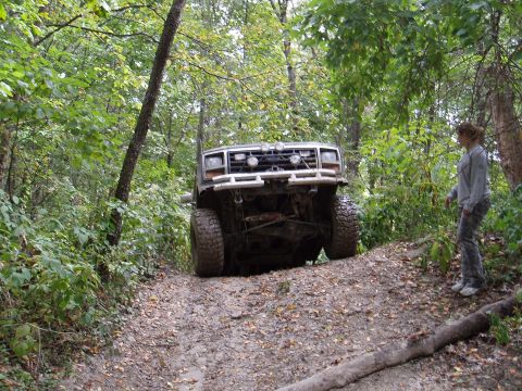1979 Ford Bronco - A Ford Bronco navigates a steep, muddy incline in a wooded area, with a person observing from the side of the trail.