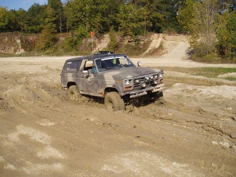 1979 Ford Bronco - A camouflaged Ford Bronco navigates a muddy off-road course, with its front-left tire deep in the mire.