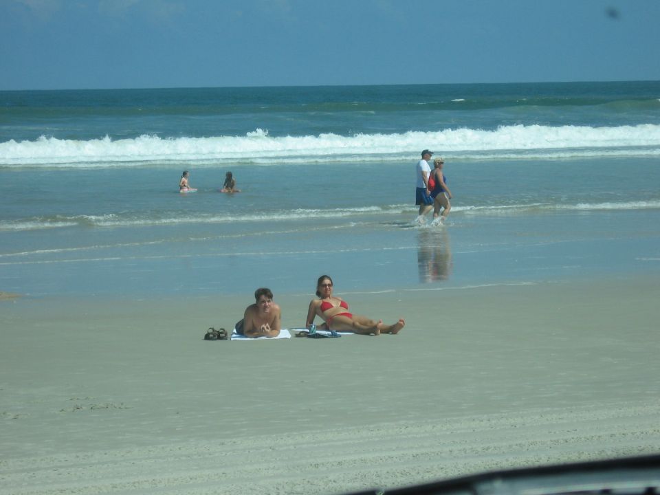 Beach scene with two people sunbathing on sand, waves in the ocean, and people walking in the water