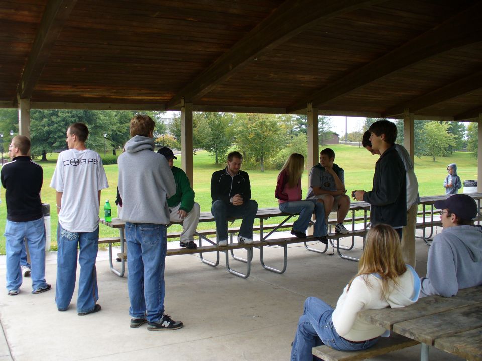 Group of people sitting and standing under a wooden picnic shelter with trees in the background