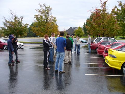 1991 Ford Probe - A group of people gather in a wet parking lot on an overcast day, surrounded by several cars.
