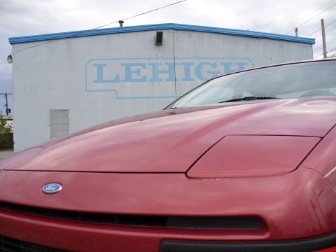 1991 Ford Probe - Low close-up across the faded maroon hood of a Ford coupe, showing the Ford oval badge on the nose panel, the closed pop-up headlamp lid an…