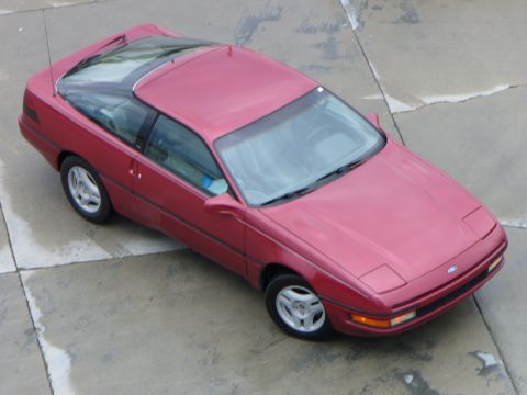 1991 Ford Probe - A red Ford Probe two-door hatchback coupe photographed from a high front-three-quarter angle on a concrete slab surface. The car has pop-up…