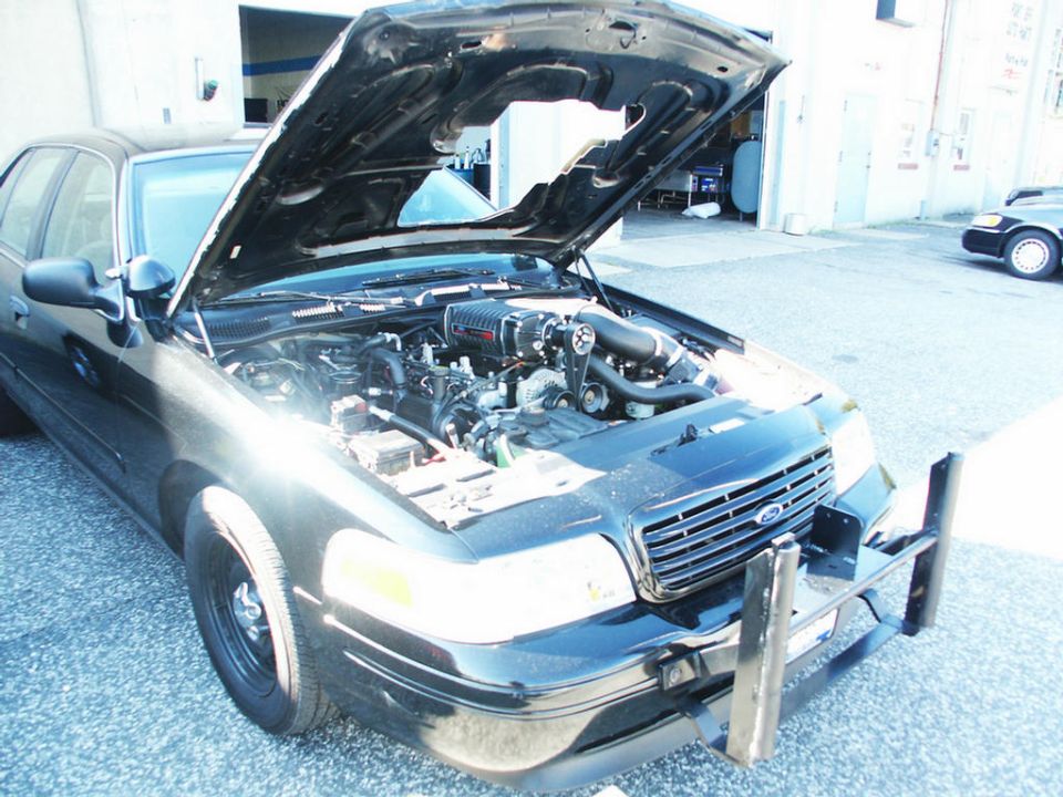 Ford Crown Victoria with hood open showing supercharger installation. Another Crown Victoria is visible in the background