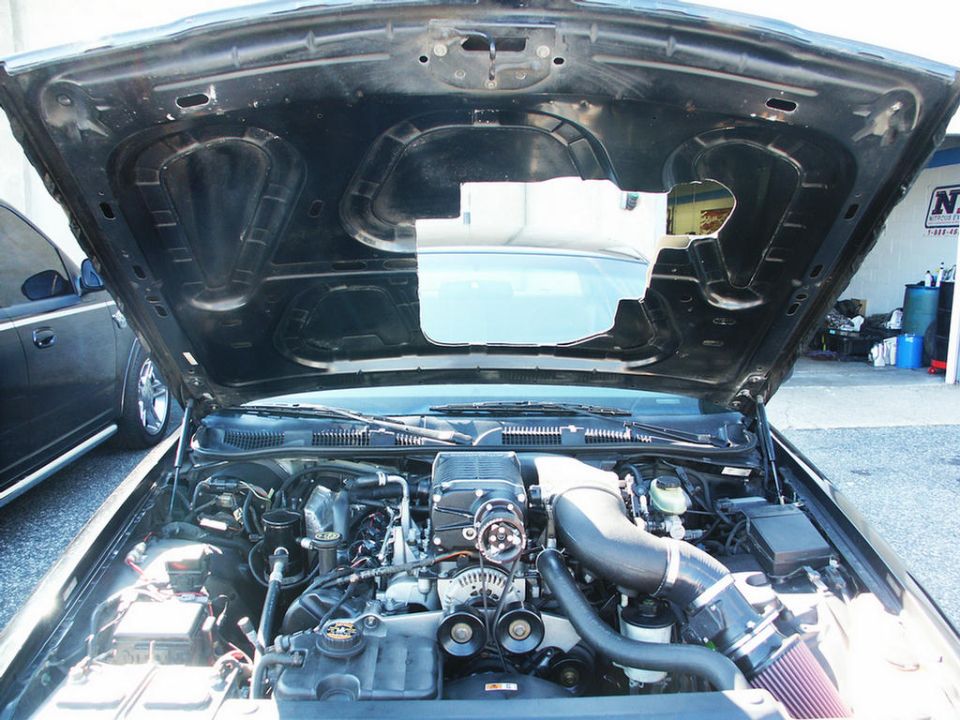 Engine bay with a black supercharger, intake, and hoses. Hood is open. Another car is visible to the left