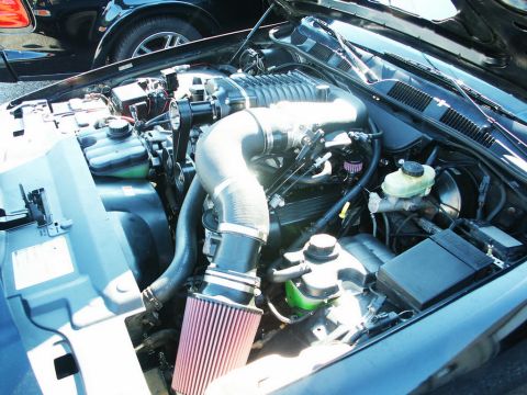 2001 Mercury Grand Marquis - The engine bay of a car is shown with a prominent supercharger and an aftermarket air filter, with another vehicle visible in the backgroun…