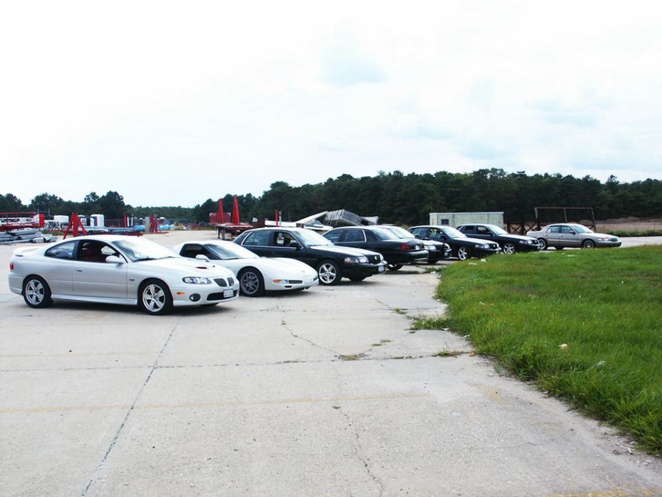 Several cars parked in a line on a paved lot, with trees and a building in the background